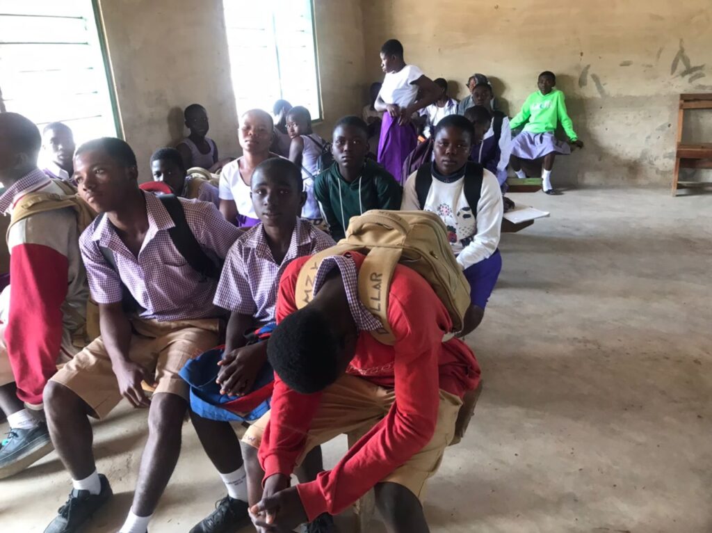 First-year students sit on benches ready for class. Some of them turn their laps into tables on which they books to take notes but others unable to do so due to overcrowding